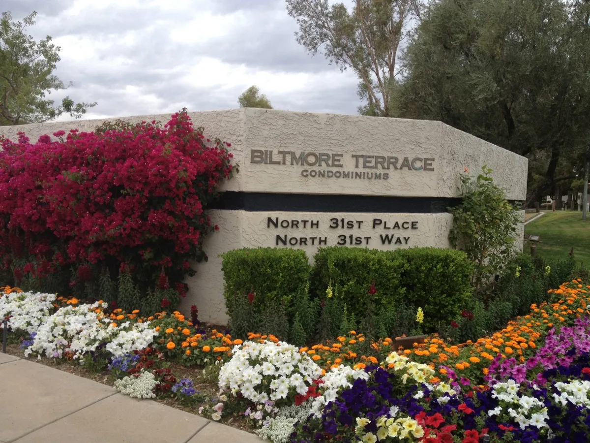 The image captures the Biltmore Terrace sign framed by vibrant flower beds and lush greenery, setting a welcoming tone for the entire community. The landscaping is meticulously maintained, and the communal garden areas are visible, suggesting opportunities for residents to stroll and enjoy the outdoors.