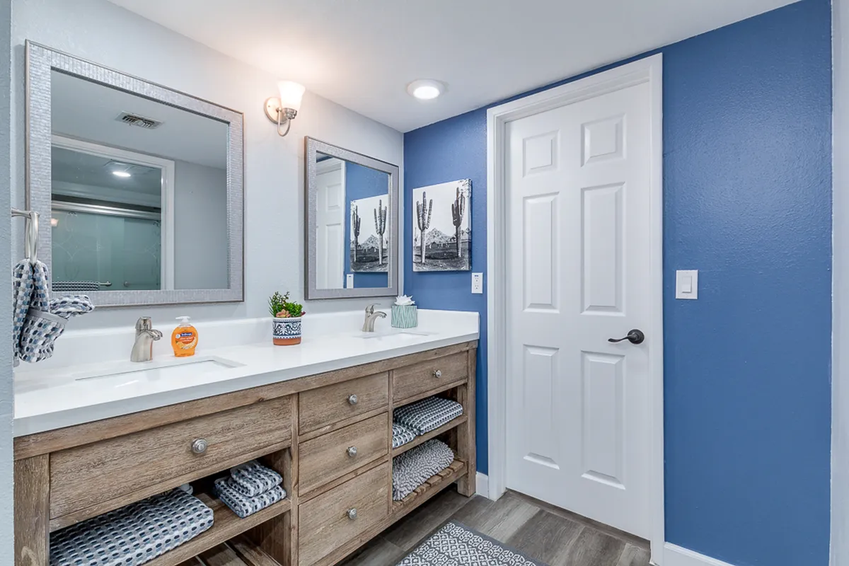 A spacious ensuite bathroom featuring a double sink vanity with large mirrors, blue accent wall, wooden cabinetry, and framed art. The space exudes a calm, refreshing ambiance ideal for self-care routines.