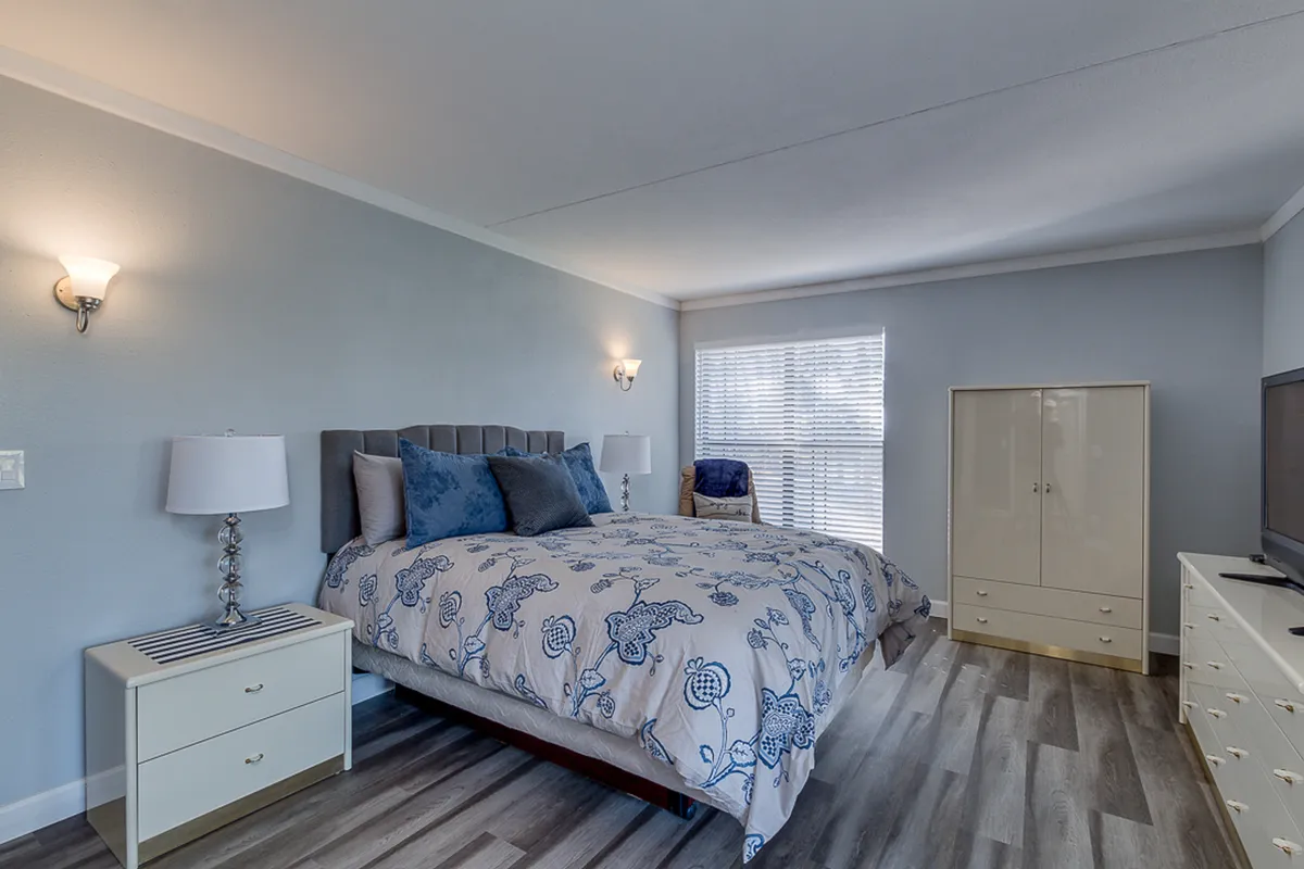 A serene bedroom with a bed adorned in decorative bedding, flanked by matching nightstands with ambient lamps. A wardrobe and in-room television are visible, creating a restful, well-appointed atmosphere ideal for unwinding after a day out.