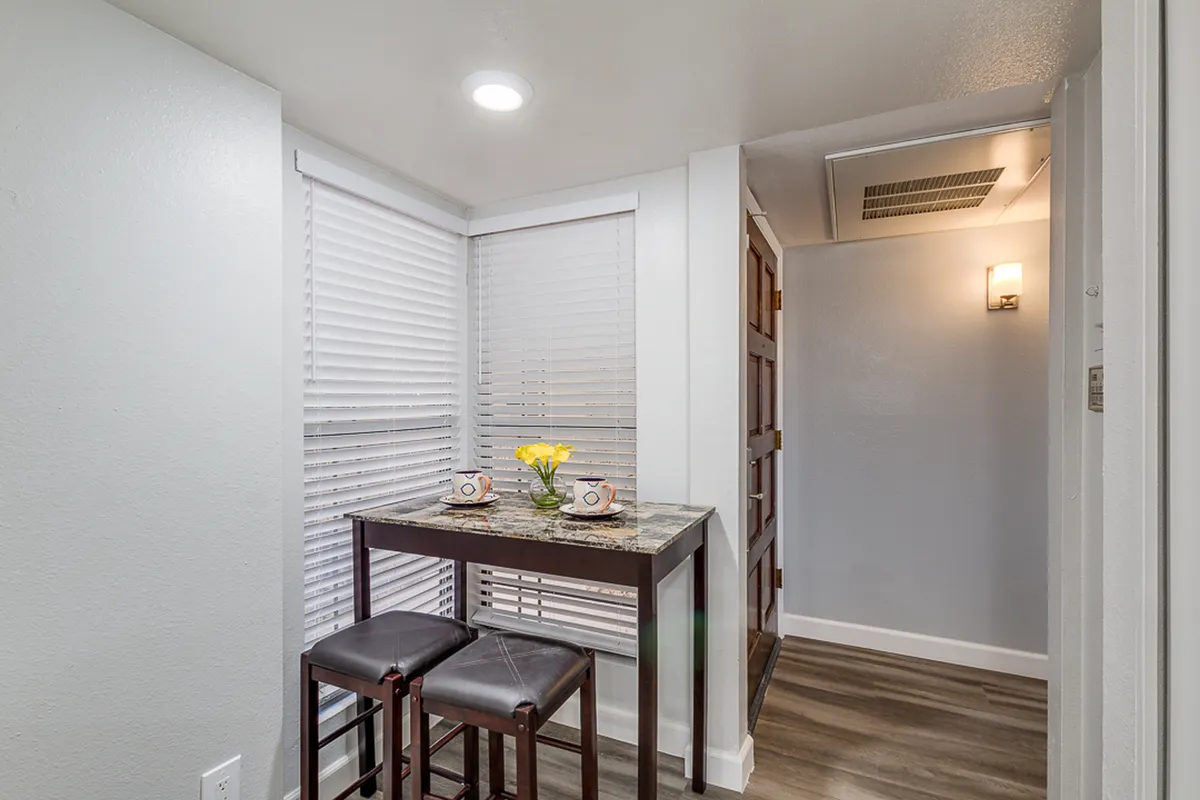 A small dining table is set with a flower vase and coffee mugs in a sunny nook, inviting cozy moments for morning coffee or casual dining. The bright light and homey touches create a welcoming atmosphere for enjoying meals.