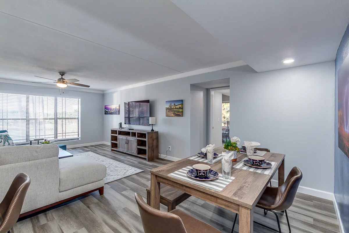 A bright, open-plan living and dining space featuring a comfortable sofa, a well-set dining table for six, and a TV unit. Warm natural light fills the area, highlighting the inviting setup for both relaxation and meals. The scene exudes coziness and is well-suited for gatherings or quiet evenings.