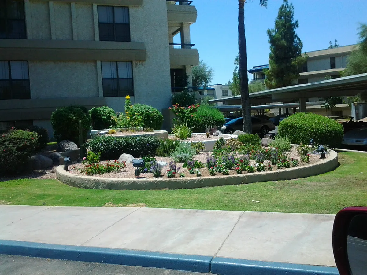 A beautifully maintained exterior flower garden is featured, with lush greenery, vibrant blooms, palm trees, and decorative stone accents. Benches are placed throughout, inviting residents to stroll or relax in the peaceful outdoor setting.